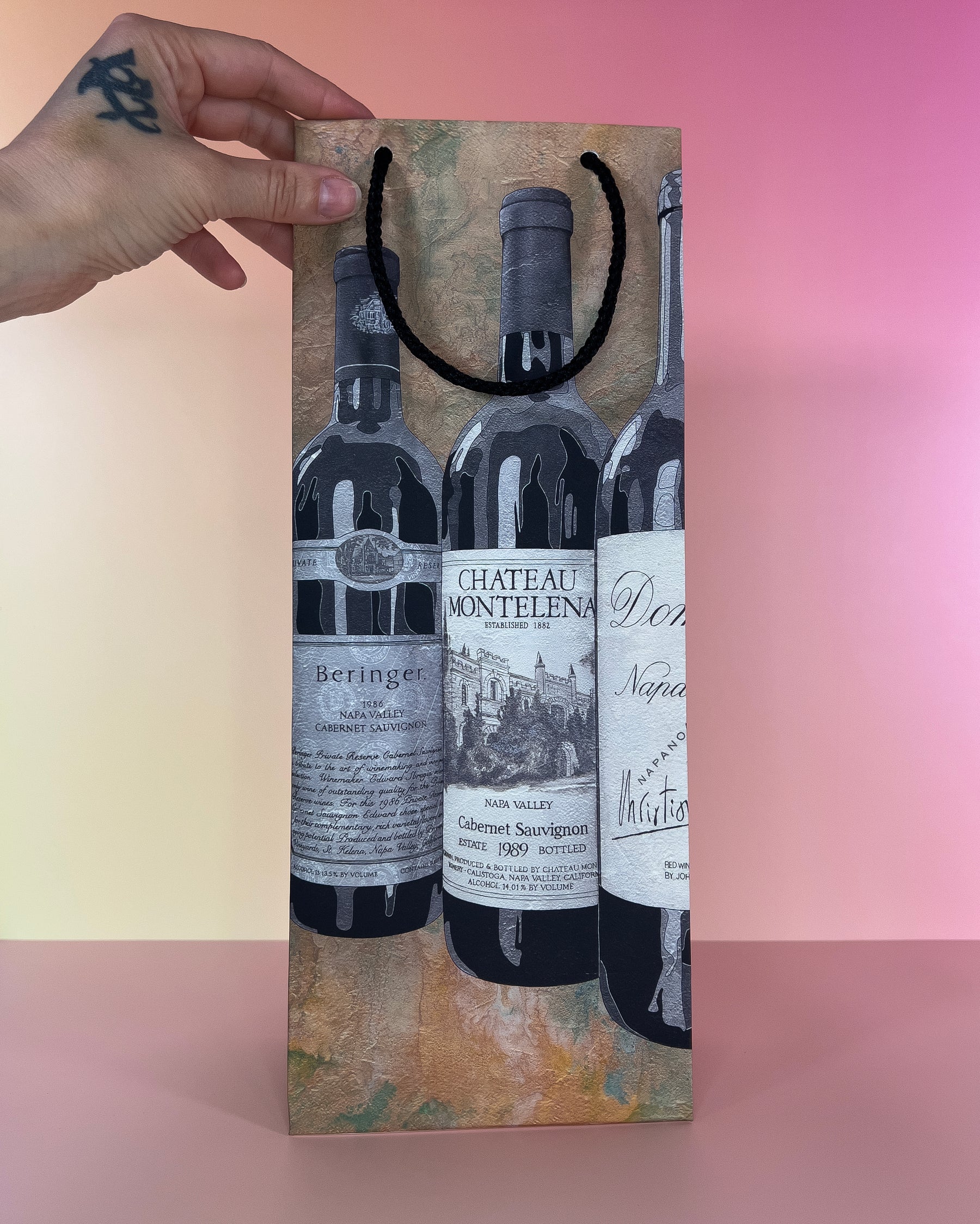 Hand holding a decorative wine gift bag with wine bottle designs on a gradient background