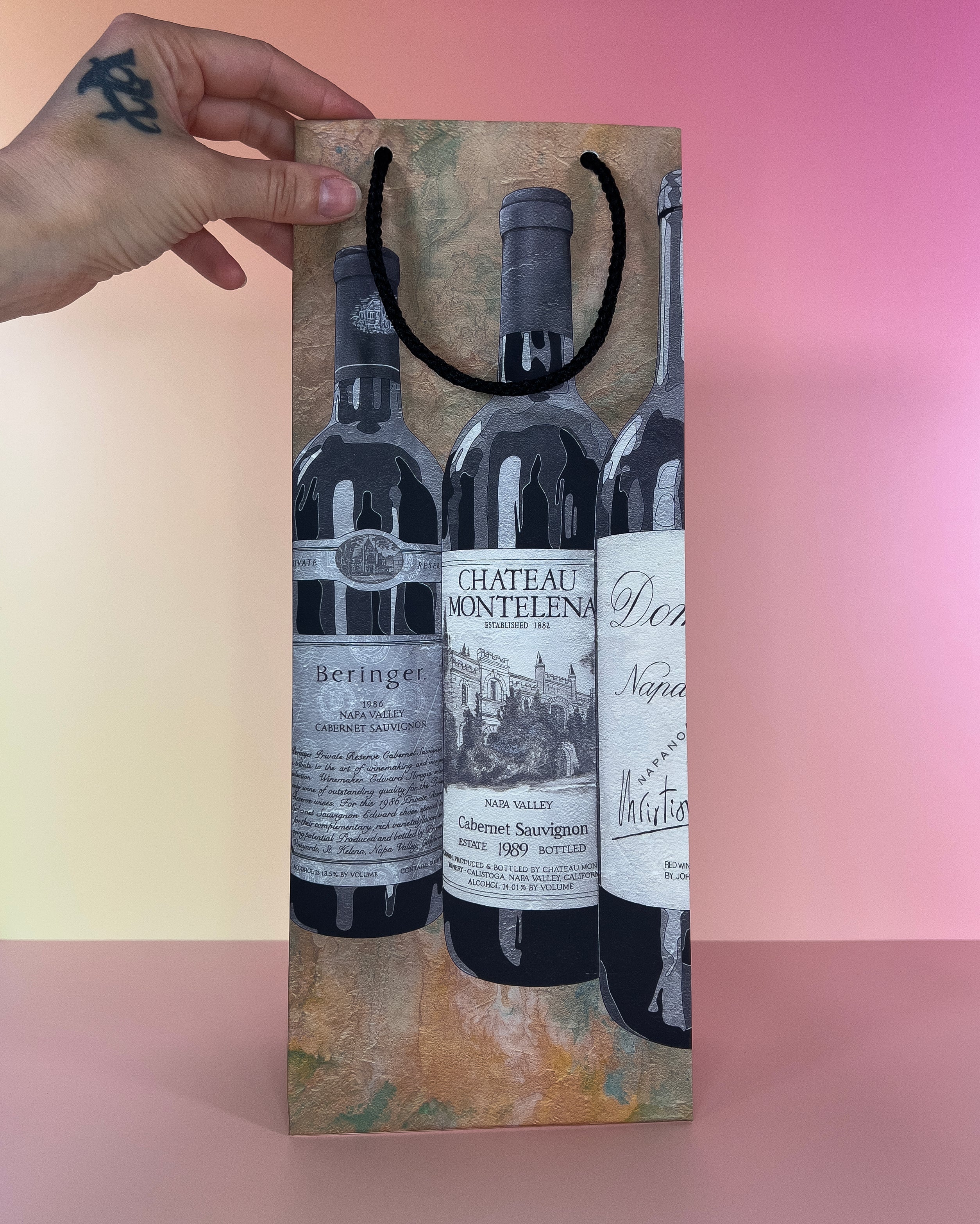 Hand holding a decorative wine gift bag with wine bottle designs on a gradient background